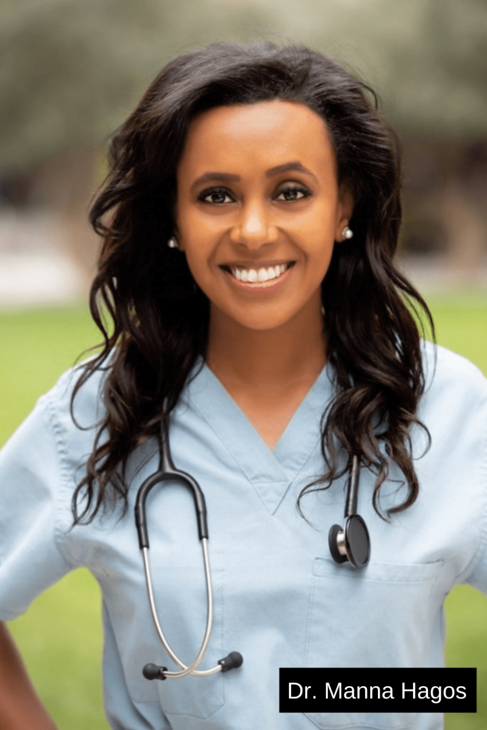 Smiling Eritrean-American female doctor wearing a stethoscope and scrubs; the doctor is standing outside.
